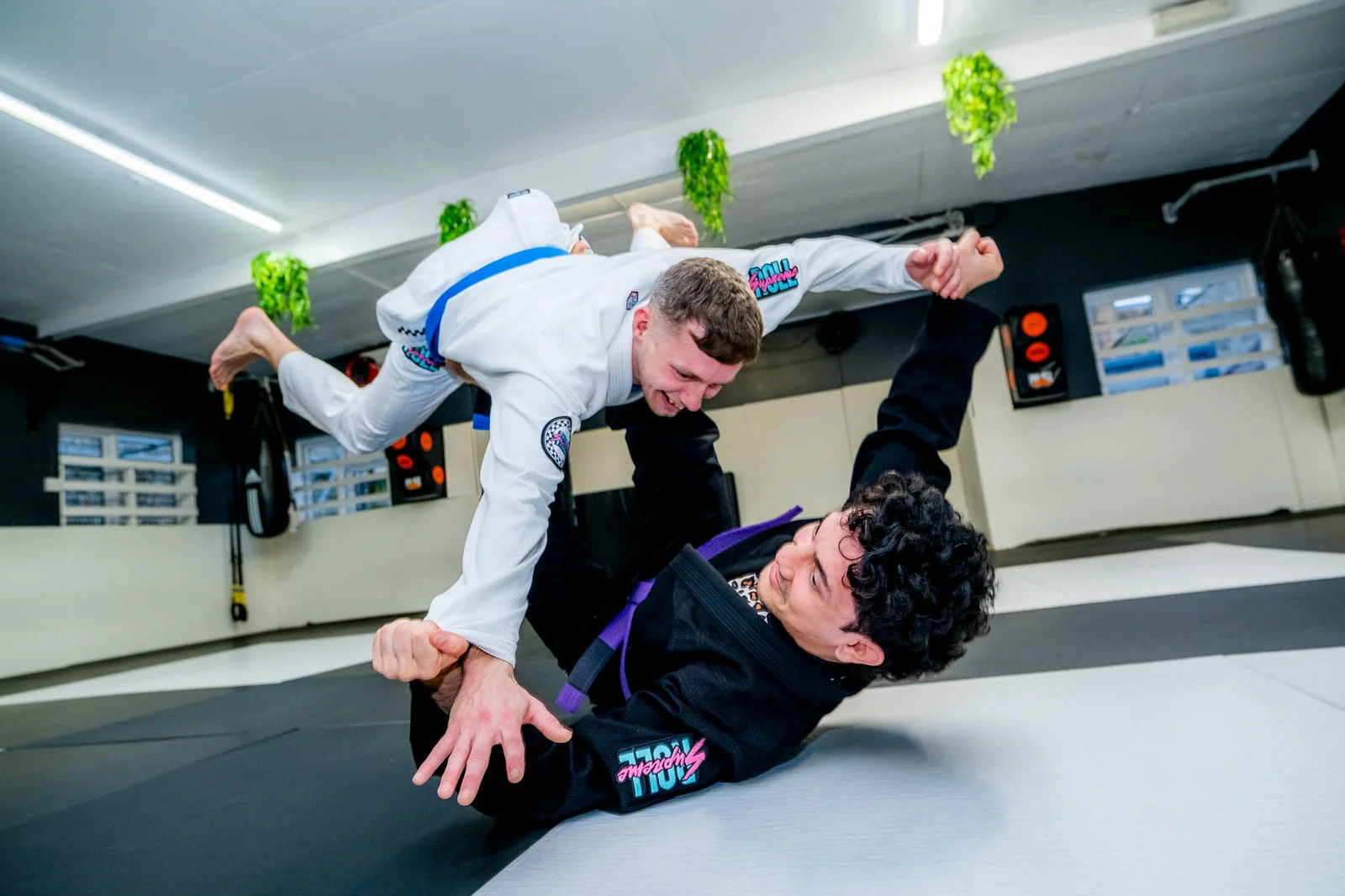 Two people training in Roll Supreme gis, one mid-air above the other, on a black and white mat.